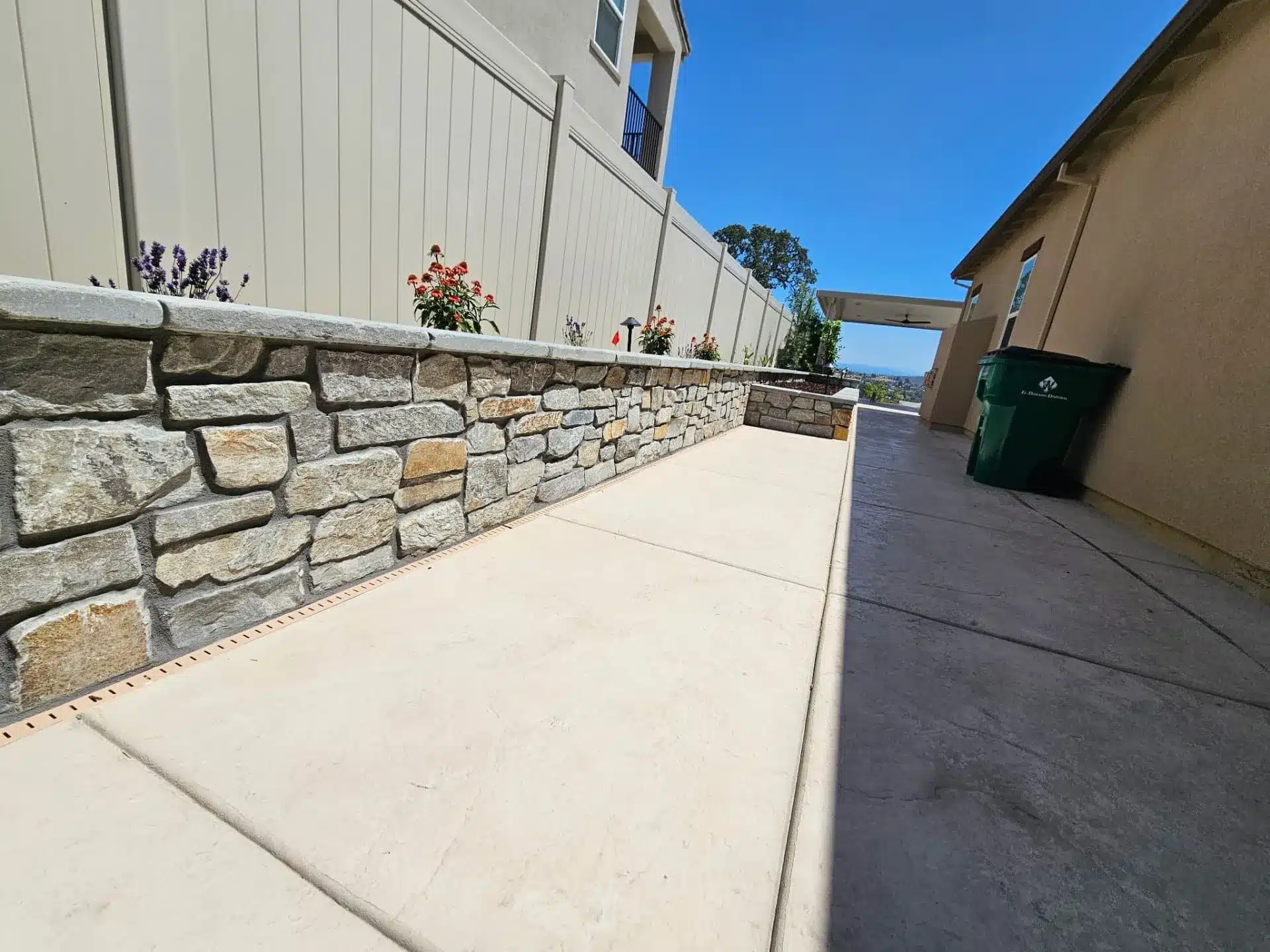 Landscaped area with stone retaining wall, concrete path, plants, and privacy fence.